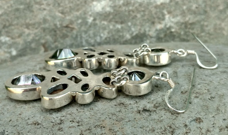 May include: A pair of silver earrings with a large, faceted, iridescent gemstone in the center of each earring. The earrings are connected to a silver chain and have a hook closure.