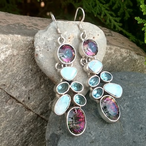 May include: A pair of silver dangle earrings with a cluster of blue and iridescent gemstones. The earrings feature a large oval-shaped iridescent gemstone at the bottom, surrounded by smaller blue and iridescent gemstones.