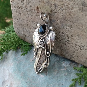 Puede incluir: Un colgante de plata con una piedra preciosa negra engastada en un dise&ntilde;o de pluma detallado.
