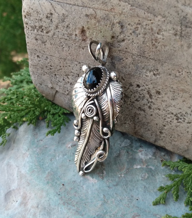 Puede incluir: Un colgante de plata con una piedra preciosa negra engastada en un dise&ntilde;o de pluma detallado.