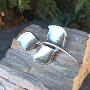 May include: A silver-toned brooch in the shape of a stylized flower or leaf. The brooch has three curved, petal-like shapes and a slender stem. The piece is displayed on a weathered, gray-brown stone surface.