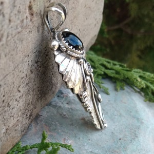 Puede incluir: Un colgante de plata con una piedra preciosa negra y un dise&ntilde;o de pluma intrincado. El colgante est&aacute; hecho de plata de ley y presenta un dise&ntilde;o de pluma detallado con una piedra preciosa de &oacute;nix negro en el centro.