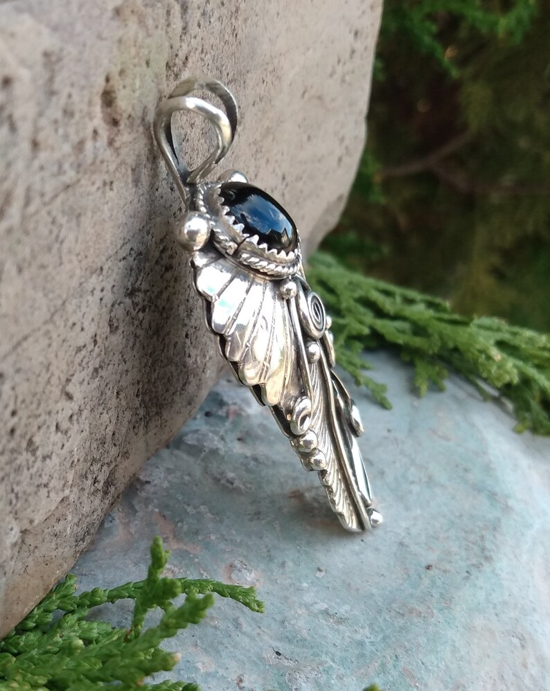 Puede incluir: Un colgante de plata con una piedra preciosa negra y un dise&ntilde;o de pluma intrincado. El colgante est&aacute; hecho de plata de ley y presenta un dise&ntilde;o de pluma detallado con una piedra preciosa de &oacute;nix negro en el centro.