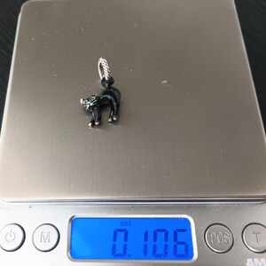 May include: A black cat charm with green eyes. The charm is on a silver-colored scale, which reads 0.106 oz. The cat is standing with its tail up and has a silver-colored loop at the top for attaching to a bracelet or necklace.