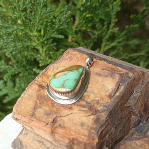 May include: A silver pendant with a teardrop-shaped turquoise stone. The stone has a light green color with brown and tan markings. The pendant is set in a silver bezel with a decorative edge and a small loop for a chain.