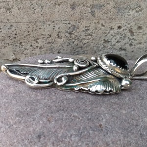 Puede incluir: Un colgante de plata con un dise&ntilde;o de pluma detallado y una piedra preciosa negra en el centro. El colgante tiene una anilla para sujetarlo a una cadena.