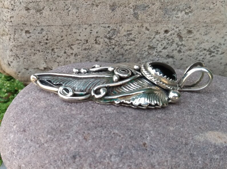 Puede incluir: Un colgante de plata con un dise&ntilde;o de pluma detallado y una piedra preciosa negra en el centro. El colgante tiene una anilla para sujetarlo a una cadena.