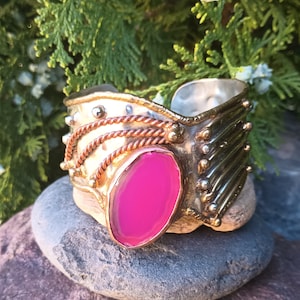 May include: A silver and brass cuff bracelet with a pink agate stone. The bracelet has a textured design and is decorated with copper wire and beads.