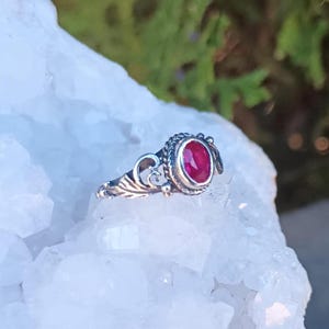 May include: A silver ring with an oval, deep red gemstone. The ring has an ornate, filigree design on the band and around the gemstone setting. The ring is displayed on a white, crystalline surface, with a blurred green background.