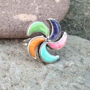 May include: A silver ring with a colorful, multi-stone design. The stones are arranged in a pinwheel pattern and are set in silver. The stones are green, orange, pink, purple, and turquoise.