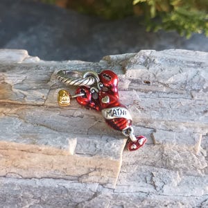 K&ouml;nnte beinhalten: Ein roter und silberner Hummer-Anh&auml;nger mit dem Wort "MAINE" darauf. Der Hummer-Anh&auml;nger hat eine goldfarbene Klaue und einen silbernen Ring zum Befestigen an einem Armband oder einer Halskette. Der Anh&auml;nger liegt auf einem Holzst&uuml;ck.