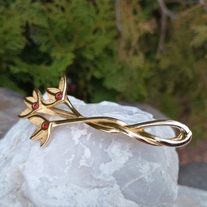 May include: Gold-toned hair clip with a twisted design and three red gemstone accents. The clip is shaped like a stylized flower.