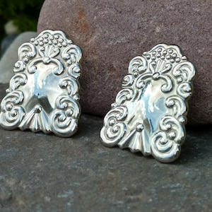 May include: Two silver earrings with a detailed, ornate design. The earrings are shaped like a rectangle with rounded corners and feature a floral pattern.