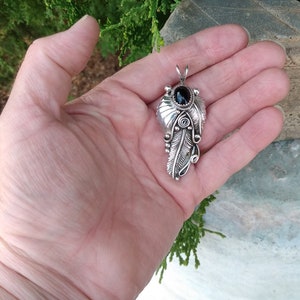Puede incluir: Un colgante de plata con una piedra preciosa negra rodeada de intrincados dise&ntilde;os de plumas.