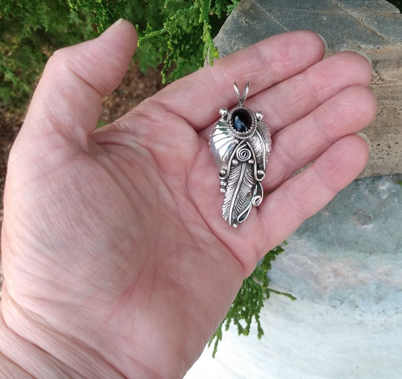 Puede incluir: Un colgante de plata con una piedra preciosa negra rodeada de intrincados dise&ntilde;os de plumas.