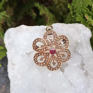 May include: A gold-toned pendant in a floral design, featuring a central red gemstone and surrounded by small, white pearls. The pendant has a loop at the top for a chain. The background is a white, crystalline surface with green foliage.