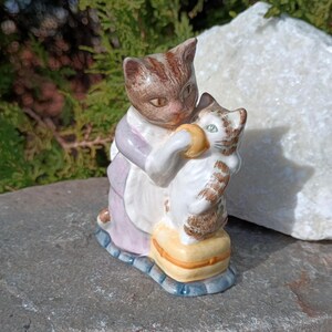 May include: A ceramic figurine depicting two cats. One cat, wearing a lavender and white outfit, embraces a smaller cat. The smaller cat is white with brown stripes and sits on a yellow cushion. The base is blue.