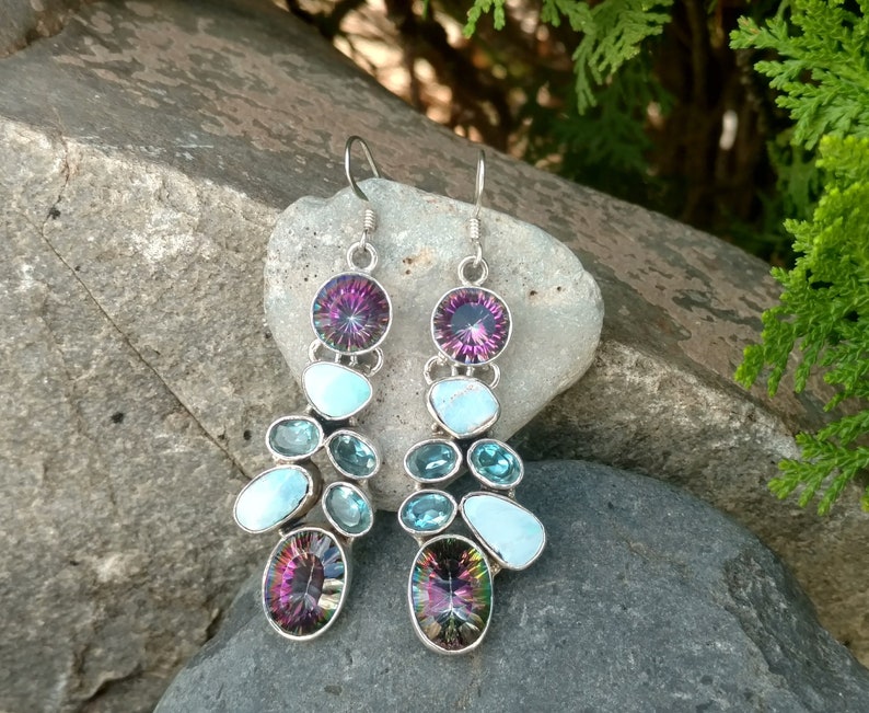 May include: A pair of dangle earrings with a silver setting and multiple gemstones. The earrings feature a large iridescent purple gemstone at the top, followed by a smaller turquoise gemstone, and then a cluster of smaller blue and turquoise gemstones.