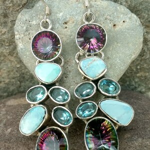 May include: A pair of dangle earrings with a mix of blue and purple gemstones. The earrings feature a large oval-shaped purple gemstone at the top, followed by a smaller round blue gemstone, and then a cluster of smaller blue and turquoise gemstones. The earrings are finished with a smaller oval-shaped purple gemstone at the bottom.