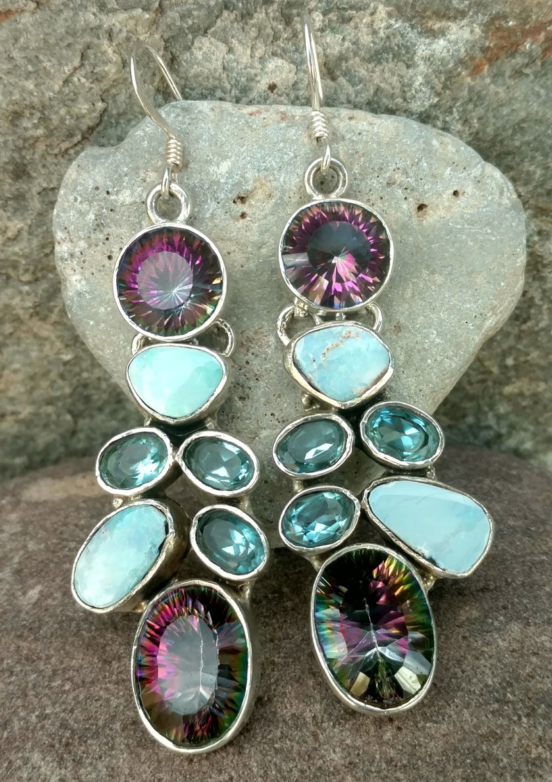May include: A pair of dangle earrings with a mix of blue and purple gemstones. The earrings feature a large oval-shaped purple gemstone at the top, followed by a smaller round blue gemstone, and then a cluster of smaller blue and turquoise gemstones. The earrings are finished with a smaller oval-shaped purple gemstone at the bottom.