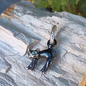 May include: A black cat charm with green eyes. The charm is made of a shiny, dark metal and has a silver-colored loop at the top. The cat is in a playful pose, with its back arched and its tail up. The charm is displayed on a gray, textured surface.