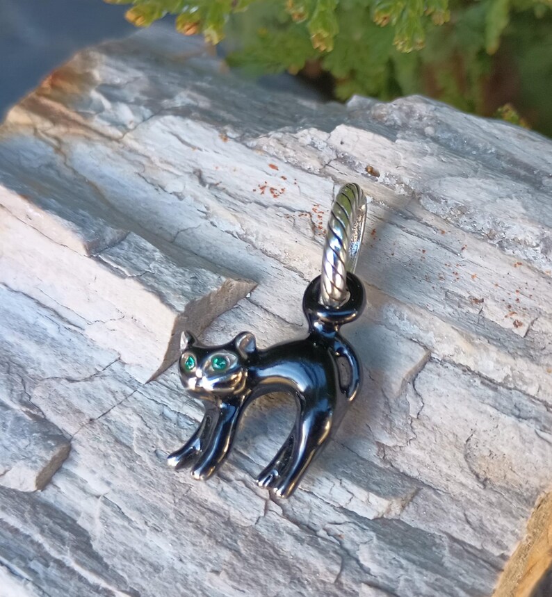 May include: A black cat charm with green eyes. The charm is made of a shiny, dark metal and has a silver-colored loop at the top. The cat is in a playful pose, with its back arched and its tail up. The charm is displayed on a gray, textured surface.