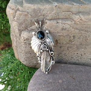 Puede incluir: Un colgante de plata con una piedra preciosa negra engastada en un dise&ntilde;o de pluma detallado.