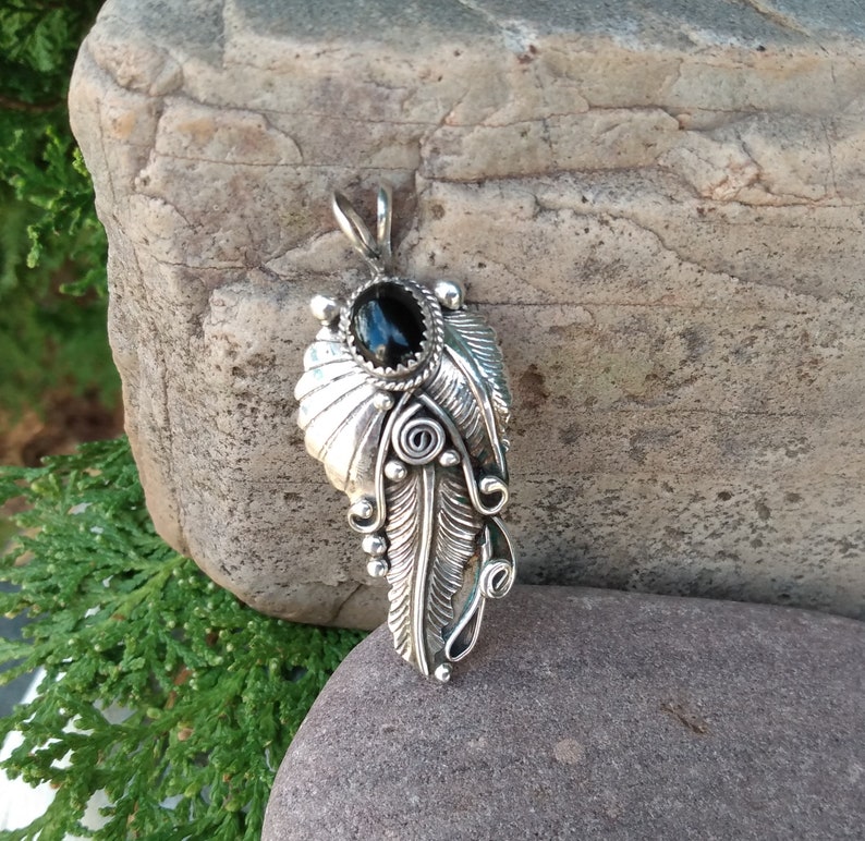 Puede incluir: Un colgante de plata con una piedra preciosa negra engastada en un dise&ntilde;o de pluma detallado.