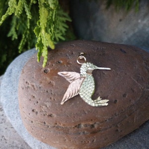 May include: A gold-toned hummingbird charm pendant with a green and pink finish. The charm is detailed with a textured surface and is perfect for jewelry making.