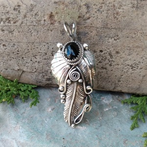 Puede incluir: Un colgante de plata con un dise&ntilde;o de pluma detallado y una piedra preciosa negra en la parte superior. El colgante est&aacute; colocado sobre un fondo de piedra gris.