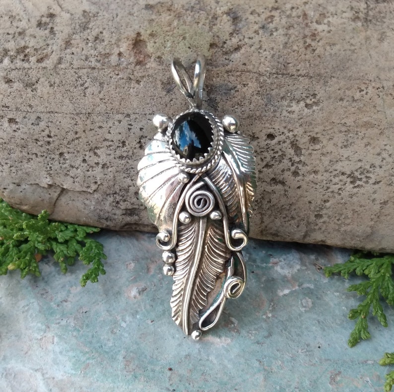 Puede incluir: Un colgante de plata con un dise&ntilde;o de pluma detallado y una piedra preciosa negra en la parte superior. El colgante est&aacute; colocado sobre un fondo de piedra gris.