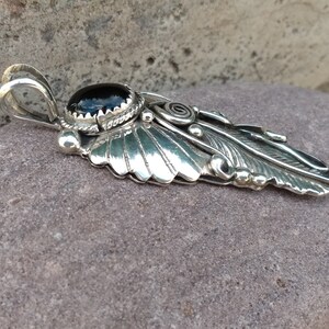 Puede incluir: Un colgante de plata con una piedra preciosa negra y un dise&ntilde;o de pluma detallado.