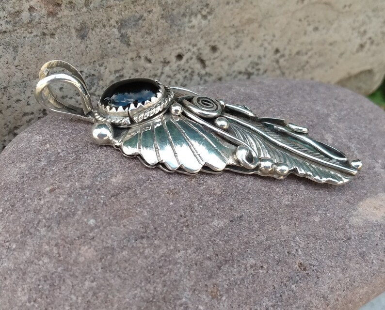 Puede incluir: Un colgante de plata con una piedra preciosa negra y un dise&ntilde;o de pluma detallado.