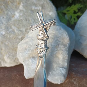May include: Silver-toned bookmark featuring a detailed windmill design. The windmill is a traditional Dutch style with a pointed roof and blades. The bookmark is a decorative piece with a functional purpose.