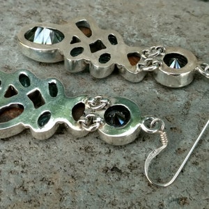 May include: A pair of silver dangle earrings with a geometric design. The earrings feature a large, faceted gemstone in the center, surrounded by smaller gemstones. The earrings are attached to a silver hook.