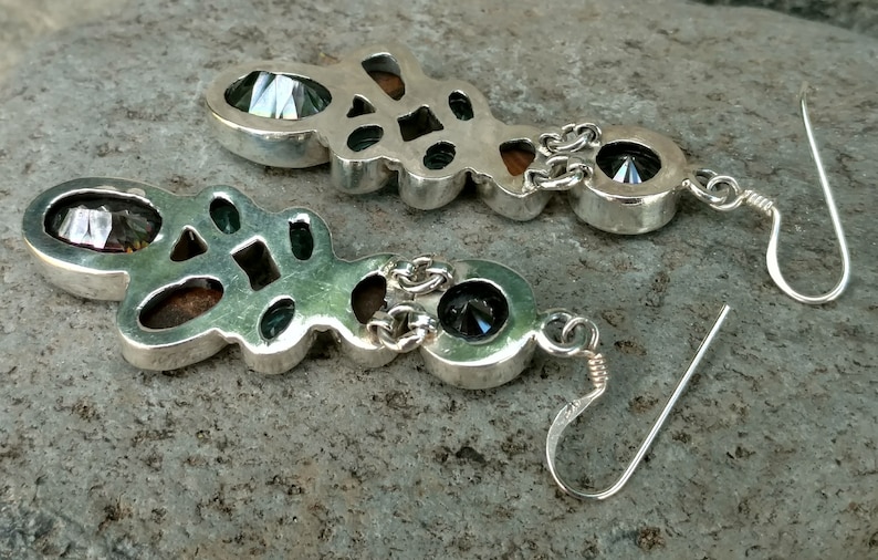 May include: A pair of silver dangle earrings with a geometric design. The earrings feature a large, faceted gemstone in the center, surrounded by smaller gemstones. The earrings are attached to a silver hook.
