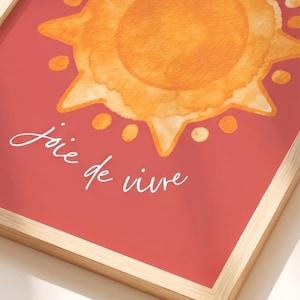 May include: A framed wall art print featuring a watercolor-style orange sun with rays and circular accents on a solid red background. The white cursive text below the sun reads joie de vivre. The print is encased in a light-colored wooden frame.