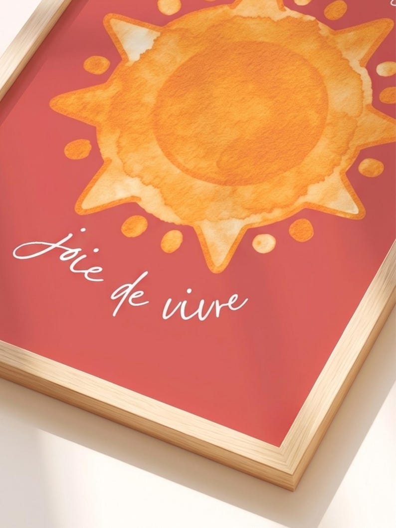 May include: A framed wall art print featuring a watercolor-style orange sun with rays and circular accents on a solid red background. The white cursive text below the sun reads joie de vivre. The print is encased in a light-colored wooden frame.
