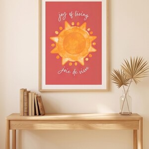 May include: A rectangular wall art print in a light wood frame featuring a watercolor-style orange sun centered on a solid red background. The text "joy of living" is printed in white above the sun, and "joie de vivre" is printed in white below.