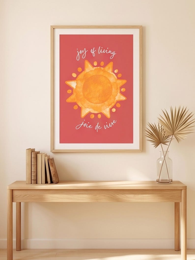 May include: A rectangular wall art print in a light wood frame featuring a watercolor-style orange sun centered on a solid red background. The text "joy of living" is printed in white above the sun, and "joie de vivre" is printed in white below.
