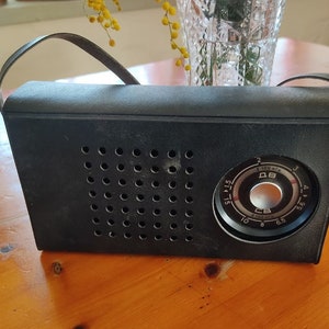Könnte beinhalten: Ein schwarzes Vintage-Transistorradio mit einem silbernen Zifferblatt und einem schwarzen Lederriemen. Das Zifferblatt hat Zahlen und Markierungen in kyrillischer Schrift.