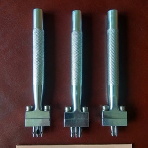 May include: Three silver leather hole punch tools with handles and three different sized punch heads. The tools are on a brown leather surface. A piece of light pink leather with three rows of holes is below the tools.