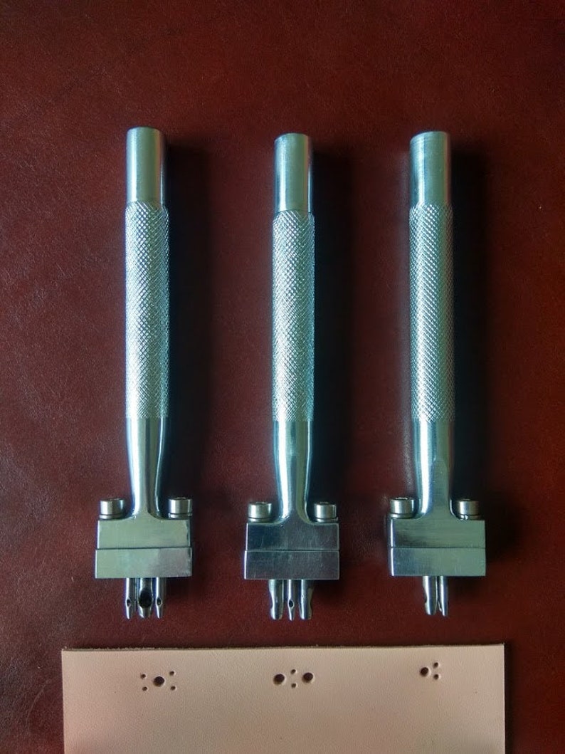May include: Three silver leather hole punch tools with handles and three different sized punch heads. The tools are on a brown leather surface. A piece of light pink leather with three rows of holes is below the tools.