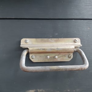 May include: Close-up of a metal handle fixed to a dark grey wooden surface. The handle is a light, aged metal with a curved grip and is secured with screws. The wooden surface has visible wood grain.