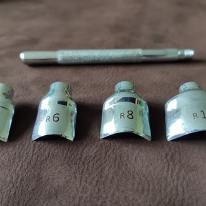 May include: Set of four silver metal tools for leather crafting. The tools are shaped like a curved blade and have different sizes marked R5, R6, R8, and R10. A silver metal handle is also included.