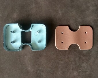 Könnte beinhalten: Ein Lederstanzwerkzeug aus Metall mit vier Löchern und einer Aussparung. Das Werkzeug liegt neben einem Stück braunem Leder mit vier gestanzten Löchern.