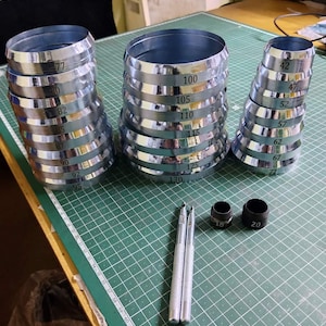 May include: A set of metal rings with different sizes, ranging from 42 to 130, stacked in three columns. The rings are silver in color and have black numbers printed on them. Two black metal punch tools are also included, one with a 16 mm diameter and the other with a 20 mm diameter.
