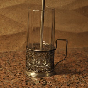 Puede incluir: Un portavasos de t&eacute; de plata con un dise&ntilde;o decorativo y un asa. El portavasos sostiene un vaso transparente con un agitador de metal en su interior.