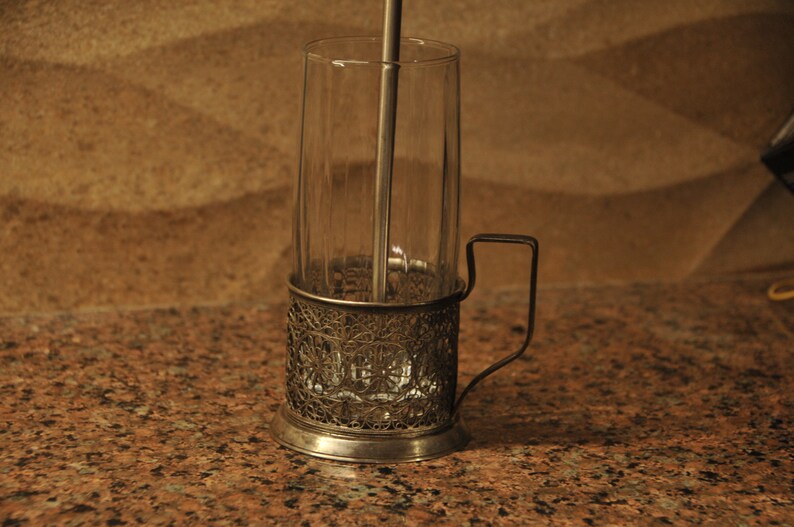 Puede incluir: Un portavasos de t&eacute; de plata con un dise&ntilde;o decorativo y un asa. El portavasos sostiene un vaso transparente con un agitador de metal en su interior.