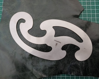 May include: A silver metal template for cutting leather with a curved design. The template is labeled "HandmadePartly" and has a small flower symbol.
